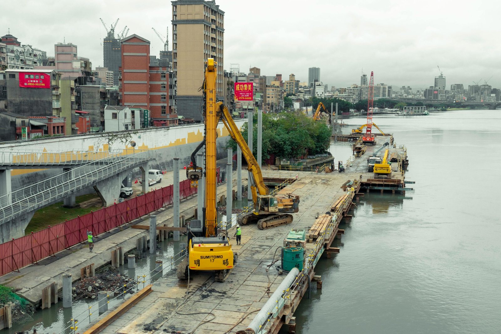 Industrial construction site by the river in Taipei with cranes and buildings in the background.